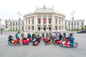 Eine Gruppe von ca. 15 Personen steht mit ihren roten Lastenrädern auf dem Vorplatz des Wiener Rathauses. Im Hintergrund ist das Burgtheater zu sehen.