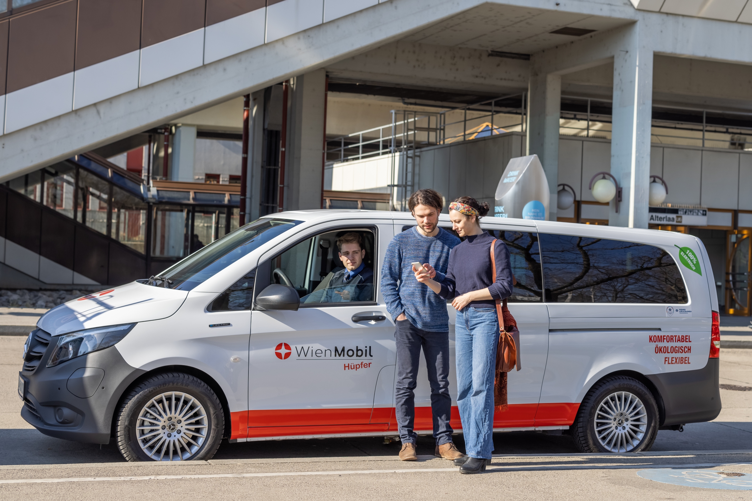 Ein großes Auto steht als WienMobil Hüpfer vor der U-Bahnstation Alterlaa. Davor stehen zwei Personen die auf ein Handy schauen, um sich womöglich eine Fahrt mit dem WienMobil Hüpfer zu buchen.